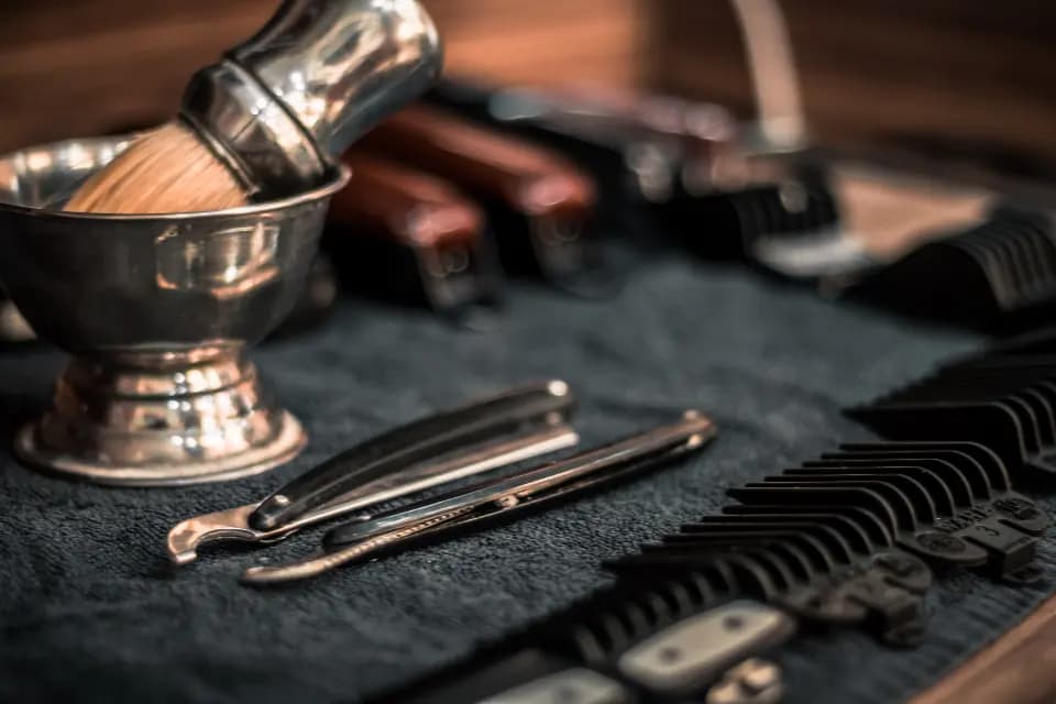 Pincel de barba, tigela de metal, navalhas e pentes de máquina sobre uma toalha escura